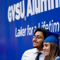 A graduate poses for a photo with a family member in front of the GVSU Alumni backdrop